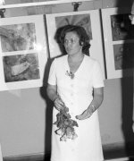 Kobieta w średnim wieku w białej sukience i z bukietem kwiatów w ręku.