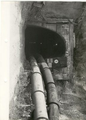 Czarnobiałe zdjęcie małego tunelu technicznego, w którym są położone rury.