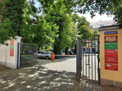 Metalowa brama wjazdowa. Za bramą szlaban. Po prawej i lewej stronie czerwone i zielone tablice z nazwami instytucji. W tle drzewa i duży budynek od strony podwórza.