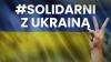 Niebiesko-żółty plakat promujący akcję #Solidarni z Ukrainą. Niebiesko-żółta flaga Ukrainy z napisem w górnej niebieskiej części #Solidarni z Ukrainą. Po prawej podniesiona ręka z dwoma palcami układającymi się w literę V.