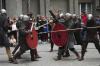 Grupa ośmiu mężczyzn ubranych w repliki średniowiecznych zbroi, z tarczami i dzidami prezentuje bitwę. Za nimi zgromadzona publiczność przygląda się i robi zdjęcia.