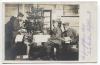 Czarnobiała pocztówka z fotografią trzech mężczyzn w starej chacie przy choince. 