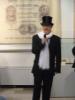 Mężczyzna w średnim wieku ubrany w garnitur, czarny kapelusz na głowie i biały szal stoi na tle ekspozycji muzealnej. Trzyma mikrofon w ręku i głosi prelekcję.