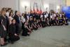Prezydent Polski wraz z Pierwszą Damą stoją wraz dużą grupą odświętnie ubranych mężczyzn i kobiet oraz młodzieży w reprezentacyjnej sali na tle biało-czerwonych flag i pozują do fotografii. Osiem osób trzyma w dłoni dyplomy.