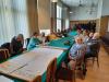 Trzynastu starszych mężczyzn i kobiet siedzi przy długim stole nakrytym zielonym obrusem w sali konferencyjnej archiwum. Na stole leży duża mapa z napisem "Brzeg Dolny". Seniorzy słuchają wykładu.