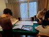 Trzy studentki stoją przy stole nakrytym zielonym obrusem w sali konferencyjnej archiwum i przyglądają się starym dokumentom. Na pierwszym planie album z czarno-białymi fotografiami Wrocławia, stara mapa oraz duże okno na ścianie za stołem.
